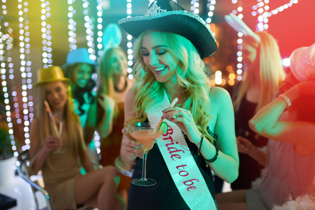 a bride to be smiles and looks down at her drink while wearing a black cowboy hat and a bride to be sash
