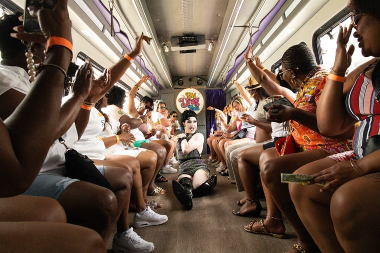 a drag queen slides across the runway floor while people cheer her on during a corporate party bus tour