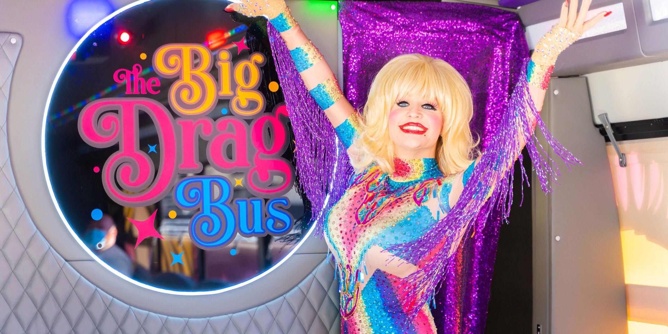 a dolly parton drag queen poses in front of the big drag bus logo