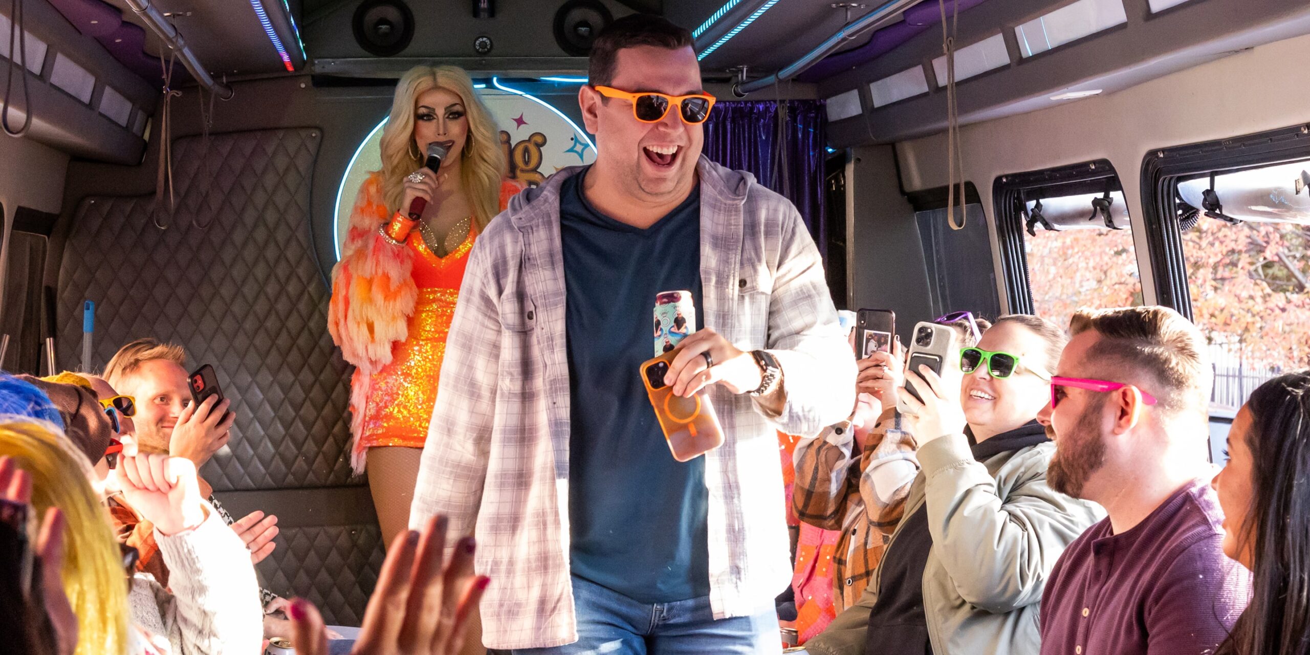 a man smiles and walks down the runway while people cheer him on and a drag queen sings behind him